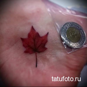 Фото тату на ладони: крошечный красный кленовый лист, расположенный на коже. Минималистичный рисунок.