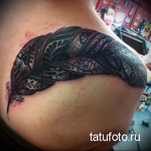 Фото тату: большое черное перо, украшенное сложным орнаментом, расположено на плече. Детализированный рисунок татуировки в стиле графика.