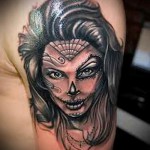 Фото тату: портрет девушки с элементами сахарной черепушки (Santa Muerte) на плече, выполненный в черно-белой гамме с яркими акцентами.