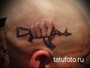 Фото тату: черно-серая татуировка на голове с изображением руки, держащей автомат, выполненная в реалистичном стиле.