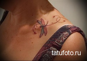 Фотография татуировки стрекозы на плече, украшенной тонкими завитушками. Рисунок выполнен в нежных розовых и серых тонах.