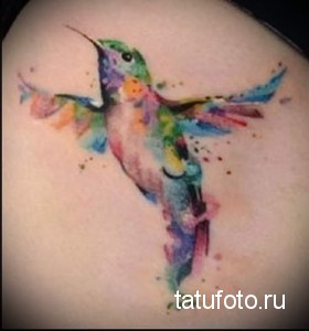Фото яркой акварельной татуировки колибри на плече. Изображение выполнено в разноцветных тонах, создающих эффект радуги.