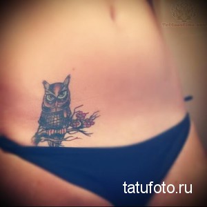 Фотография небольшой татуировки совы, сидящей на ветке с цветами, на боку девушки. Черно-серый рисунок с детальной проработкой оперения и лепестков.