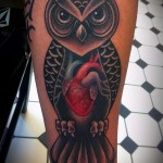 Фото тату совы на руке с сердцем в центре груди. Черно-серый рисунок с ярким красным сердцем.