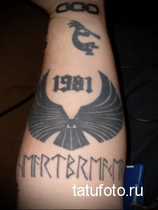 Фото тату ворона, рунической надписи и цифры 1981 на предплечье. Рисунок выполнен в черном цвете с использованием четких линий и геометрических форм.