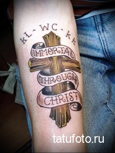 Фото тату креста с надписью 'IMMORTAL THROUGH CHRIST' на предплечье. Рисунок выполнен в коричневых и черных тонах, с использованием штриховки и теней.