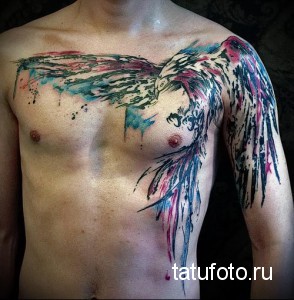 Фото тату акварельного феникса на груди и плече. Яркие розовые, голубые и черные оттенки, брызги краски, создающие эффект огня и возрождения.