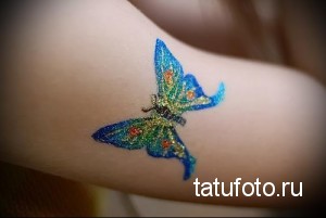 Фотография глиттер-тату с изображением бабочки на руке. Используются синие, зеленые и золотые блестки. Красивый и нежный дизайн.