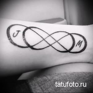 Фото татуировки на запястье в виде символа бесконечности, внутри которого расположены буквы 'J' и 'M'. Черно-белое изображение, минималистичный стиль.