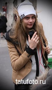 Фотография модели в белой шапке с надписью и бежевой куртке, идущей по улице с телефоном в руке, демонстрирующая современный городской стиль.