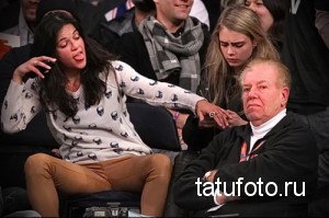 Фотография девушки, сидящей на трибуне с высунутым языком. На ней белая кофта с принтом черепов и коричневые брюки. Рядом с ней сидит мужчина и другая девушка.