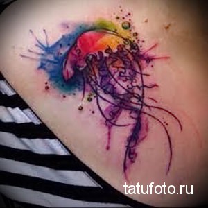 Фото акварельной татуировки медузы на плече. Рисунок выполнен в ярких цветах, с брызгами краски и плавными линиями.
