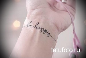 Фото тату: надпись 'Be Happy' изящным шрифтом на запястье. Минималистичная татуировка с черными чернилами, символ позитива и оптимизма.
