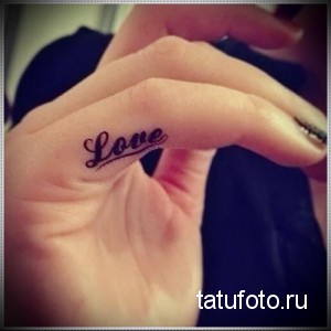 Фотография маленькой татуировки надписи 'Love' на фаланге пальца. Черно-белый рисунок татуировки, выполненный каллиграфическим шрифтом.