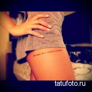 Фото тату: Надпись "Curiosity often leads to trouble" на женском бедре, выполненная элегантным шрифтом. Стильная и минималистичная татуировка.