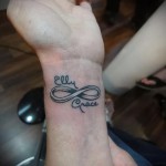 Фото тату на запястье: знак бесконечности с именами 'Elly' и 'Grace' внутри. Черно-серый рисунок татуировки, выполненный в каллиграфическом стиле.