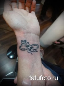 Фото тату на запястье: знак бесконечности с именами 'Elly' и 'Grace' внутри. Черно-серый рисунок татуировки, выполненный в каллиграфическом стиле.