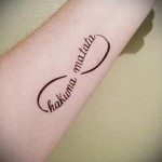 Фото тату бесконечность с надписью 'Hakuna Matata' на предплечье. Черный контур, минималистичный дизайн, популярный эскиз татуировки.