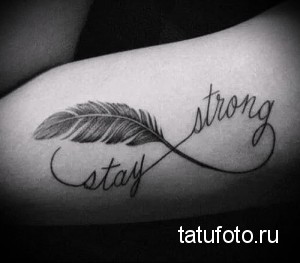 Фото тату бесконечность с пером и надписью 'Stay Strong' на предплечье. Черно-белый рисунок татуировки, выполненный в графичном стиле, с акцентом на детали пера.