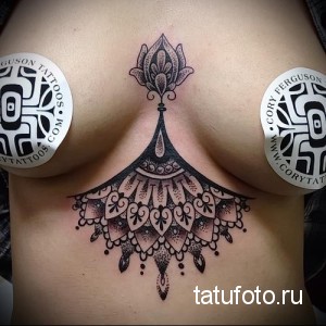 Фото татуировки под грудью женщины, выполненной в технике дотворк. Изображена сложная мандала с множеством мелких деталей и геометрических узоров.