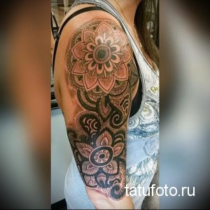 Фото тату на плече: мандала, окруженная цветами и растительными узорами. Татуировка выполнена в нежных розовых и черных тонах.