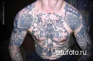 Фото тату на груди и руках: сложная композиция с крестом, ангелами, звездами и религиозными надписями. Детализированный рисунок татуировки в черно-серых тонах.