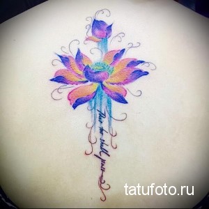 Фотография цветной татуировки лотоса на спине девушки. Акварельные переходы цветов, надпись и изящный дизайн.