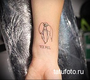 Фото тату небольшого ангела с крыльями и надписью 'VEML' на запястье. Черный контур, простой дизайн.