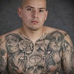 Фото тату на груди и плечах мужчины: сложный рисунок с черепами, розами, надписями и другими символами. Черно-белая татуировка с высокой детализацией.