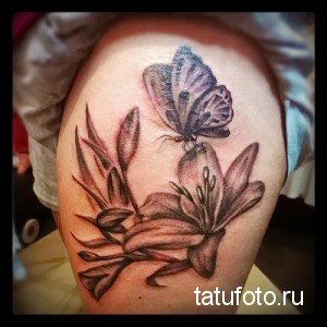 Фото татуировки бабочки, сидящей на цветке лилии, выполненной в реалистичном стиле. Детализированный рисунок с использованием теней и световых эффектов.