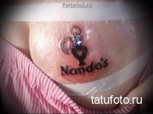 Фото тату логотипа ресторана Nando's на груди. Черно-красный рисунок татуировки с изображением петуха и надписью 'Nando's'.
