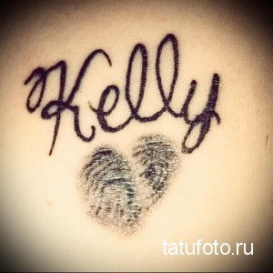Фотография татуировки с именем 'Kelly' и отпечатком пальца в форме сердца, расположенной на коже. Рисунок выполнен черными чернилами.