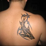 Фото тату: стилизованная татуировка акулы в стиле трайбл на спине, выполненная в черных тонах с использованием геометрических узоров и синих акцентов.