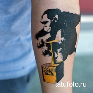 Фото татуировки обезьяны, пьющей из бочки с ядом. Рисунок выполнен в стиле стрит-арт с использованием черного и желтого цветов. Подчеркивает протест и бунтарство.