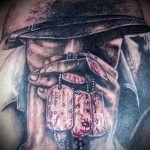Черно-серое фото тату на плече с изображением руки, держащей цепочку с жетонами и шляпой. Детализированный рисунок, реалистичный стиль.