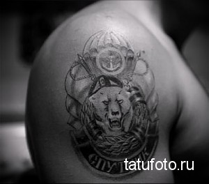Фото тату на плече с изображением геральдического щита, головы волка и надписи "Служить Родине". Выполнено в черно-серых тонах.