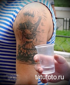 Фото тату воина с автоматом и саблей на фоне горящего города на плече. Черно-серая татуировка в реалистичном стиле.