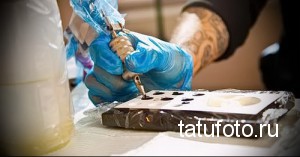 Фотография процесса татуировки, где мастер в голубых перчатках наносит рисунок на кожу клиента, используя тату-машинку и трафарет.