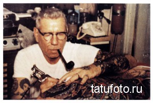 Фотография старого тату-мастера за работой в своей мастерской. Видны инструменты для татуирования и эскизы на стенах.