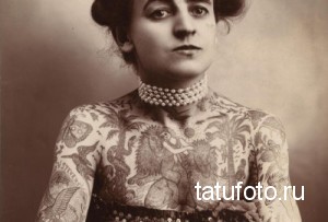 Старинное фото женщины начала XX века, покрытой татуировками на руках, шее и теле. Изображены цветы, животные и другие декоративные элементы.