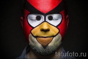 Фото тату-макияжа в виде персонажа Angry Bird на лице мужчины с бородой. Красный, желтый и черный цвета.