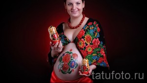 Фото тату: Боди-арт на животе беременной женщины с изображением матрешек и русских цветов. Красивый рисунок татуировки.