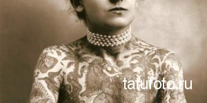 Старинное фото женщины с густыми татуировками, покрывающими ее руки, шею и грудь. Она носит жемчужное ожерелье и смотрит прямо в камеру.