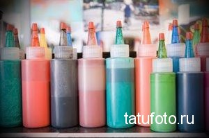 Фотография ряда бутылочек с разноцветными пигментами для татуировок, расположенных на полке. Включает оттенки красного, оранжевого, желтого, зеленого, синего и серого.