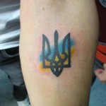 Фото минималистичной татуировки с изображением украинского герба - Тризуба, выполненного в черном цвете с добавлением акварельных синих и желтых мазков.