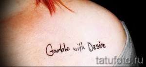 Фото татуировки с надписью 'Gamble with Desire' на плече. Рукописный шрифт, черные чернила, немного небрежный стиль.