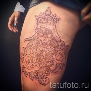 Фото тату Калавера в короне с цветами и костями на бедре. Детализированный рисунок татуировки в стиле графика.