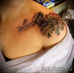 Фото татуировки с ромашкой, бабочкой и надписью 'Beloved' на плече. Использованы нежные оттенки розового и серого.