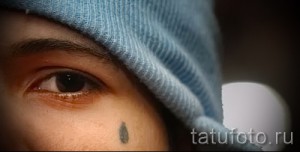 Фото тату маленькой слезы под глазом мужчины. Минималистичный рисунок татуировки, выполненный черным цветом, символизирующий печаль или потерю.