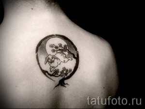 Фото тату на лопатке: черно-белый рисунок зайца, сидящего под цветущей веткой сакуры, заключенного в круг.